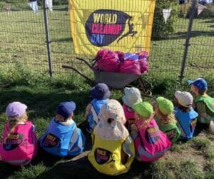 Kleinkinder mit Sonnenhüten und World-Cleanup-Day-Westen sitzen von hinten fotografiert im Gras vor einer Schubkarre voller pinker Müllsäcke und einer gelben World-Cleanup-Day-Fahne.
