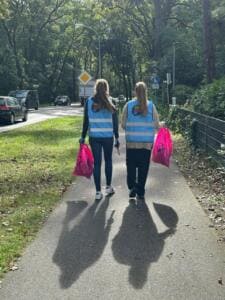 Zwei Frauen in blauen World-Cleanup-Day-Warnwesten gehen mit pinken Müllsäcken einen sonnigen Radweg entlang, von hinten fotografiert.