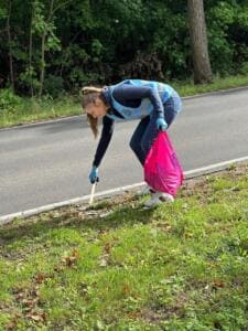 Eine junge Frau in blauer Warnweste hebt mit einer Greifzange Müll vom Grünstreifen an einer Landstraße auf und hält einen pinken Sack.