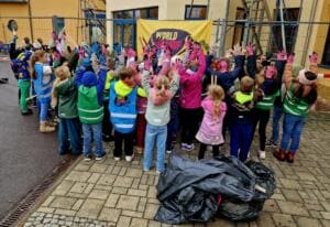 Viele Grundschulkinder in bunten Warnwesten recken vor einem gelben World-Cleanup-Day-Banner ihre Hände mit Arbeitshandschuhen in die Luft, davor gefüllte Müllsäcke.
