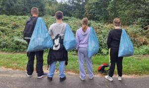 Vier Jugendliche stehen von hinten fotografiert mit vollen hellblauen Müllsäcken über der Schulter an einem Weg.