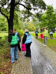 Mehrere Personen in Warnwesten sammeln mit pinken Säcken entlang eines nassen Parkwegs zwischen Bäumen.