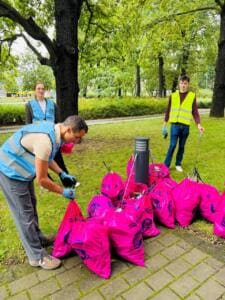 Ein Mann in blauer Warnweste bindet im Park einen von vielen gefüllten pinken World-Cleanup-Day-Säcken zu, dahinter zwei weitere Helfende.