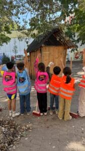 Kinder in pinken, blauen und orangefarbenen Warnwesten stehen mit Besen auf einem Hof vor einem Holzhäuschen und heben die Hände, von hinten fotografiert.