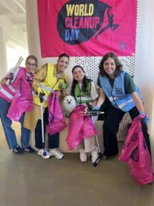 Vier lachende Frauen in bunten Warnwesten mit pinken Müllsäcken und Greifzangen posieren mit einem weißen Hund vor einer World-Cleanup-Day-Fahne im Büro.