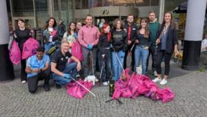 Eine Gruppe Erwachsener mit pinken Müllsäcken und Greifzangen posiert vor dem Eingang eines Einkaufszentrums.