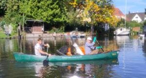 Zwei Männer paddeln ein grünes Kanu, das mit gesammeltem Müll beladen ist, über einen Fluss vor Ufergrundstücken mit Booten.