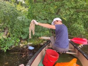 Eine Frau sitzt in einem Kanu am bewachsenen Ufer und zieht einen verschmutzten Plastikbeutel aus dem Wasser; im Boot stehen ein roter Eimer und ein orangefarbener Sack.