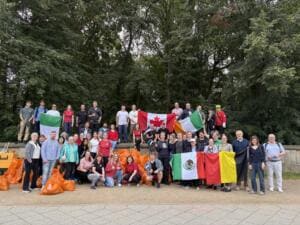 Eine große Gruppe Erwachsener steht mit Länderflaggen aus Kanada, Irland, Mexiko und Belgien hinter vielen orangefarbenen Müllsäcken am Parkrand.