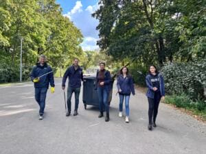 Fünf Erwachsene in dunkelblauen Jacken laufen mit Greifzangen und einer Mülltonne eine von Bäumen gesäumte Straße entlang.