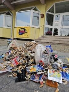 Ein Haufen gesammelter Müll mit vielen Zigarettenstummeln, Trinkpäckchen und Plastikverpackungen liegt vor einem Gebäudeeingang mit gelbem World-Cleanup-Day-Banner.