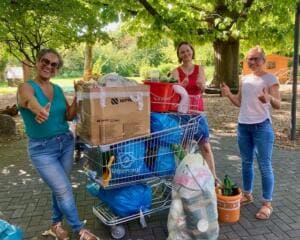 Drei Frauen zeigen lachend den Daumen hoch neben einem Einkaufswagen voller Müllsäcke, Kartons und Flaschen.
