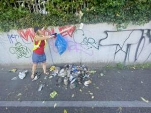 Eine Frau in gelber Warnweste sammelt mit Greifzange Müll an einer mit Graffiti besprühten Mauer, davor ein Haufen Flaschen und Dosen.