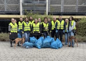 Eine Gruppe Erwachsener in gelben Warnwesten steht mit zwei Hunden hinter vielen gefüllten blauen Müllsäcken vor einem Parkhaus.