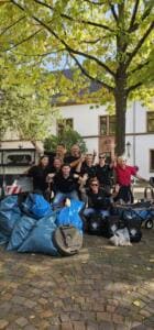 Eine Gruppe Erwachsener in schwarzen T-Shirts jubelt vor einem historischen Gebäude hinter vielen blauen Müllsäcken und Eimern.