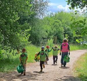 Kleine Kinder in World-Cleanup-Day-Westen laufen mit grünen Eimern hinter einer Erzieherin einen Waldweg entlang, von hinten fotografiert.