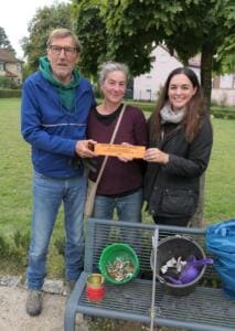 Drei Erwachsene halten gemeinsam ein oranges Schild, vor ihnen auf einer Bank Eimer mit gesammelten Zigarettenstummeln und eine Blechdose.