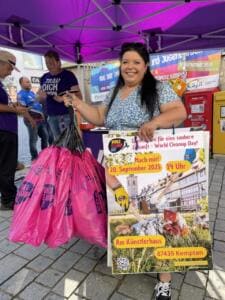 Eine lächelnde Frau unter einem Pavillon hält ein Plakat zum World Cleanup Day am 20. September 2025 in Kempten und mehrere pinke Müllsäcke.