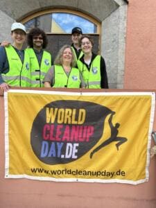 Fünf Erwachsene in gelben Warnwesten stehen lachend hinter einem gelben World-Cleanup-Day-Banner.