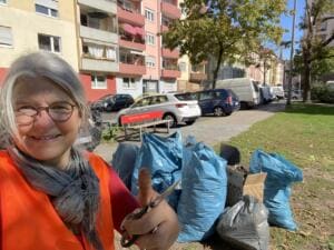 Selfie einer Frau in orangefarbener Warnweste mit Daumen hoch, hinter ihr am Straßenrand viele gefüllte blaue Müllsäcke.