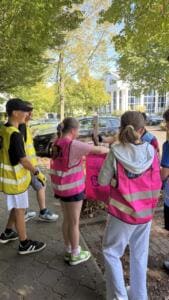 Mehrere Jugendliche in pinken und gelben Warnwesten sammeln Müll auf einem Gehweg unter Bäumen, überwiegend von hinten fotografiert.