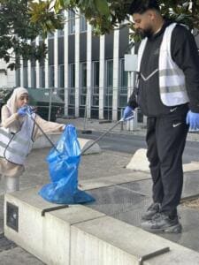 Eine Frau und ein Mann in Warnwesten sammeln mit Greifzangen Müll in einen blauen Sack auf einem Platz vor einem Bürogebäude.
