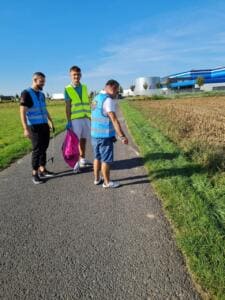 Drei junge Männer in Warnwesten mit Greifzange und pinkem Sack stehen auf einem Weg zwischen Wiese und Feld, im Hintergrund ein Gewerbegebiet.