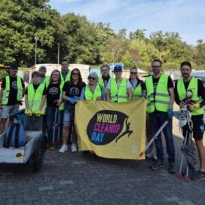 Eine Gruppe Erwachsener und ein Kind in gelben Warnwesten mit Greifzangen hält eine gelbe World-Cleanup-Day-Fahne, daneben ein Handwagen.