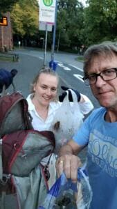 Selfie eines Mannes und einer jungen Frau, die an einer Bushaltestelle gefüllte Plastiktüten mit Müll hochhalten.