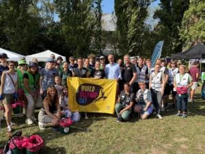 Eine große Gruppe Erwachsener und Kinder mit Westen und Eimern steht im Park vor Pavillons um eine gelbe World-Cleanup-Day-Fahne.