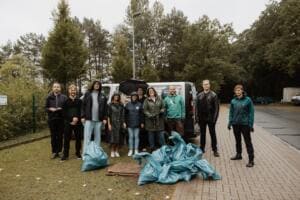 Eine Gruppe Erwachsener in Regenkleidung steht auf einem Parkplatz vor Transportern, davor gefüllte türkisfarbene Müllsäcke.