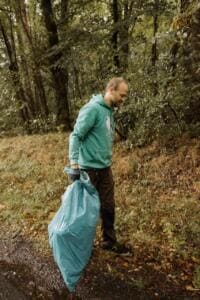 Ein Mann im grünen Kapuzenpullover trägt einen gefüllten türkisfarbenen Müllsack am Waldrand.