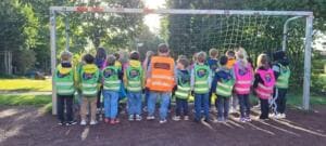 Eine Grundschulklasse in bunten World-Cleanup-Day-Westen steht von hinten fotografiert vor einem Fußballtor.