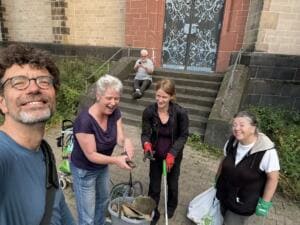 Selfie eines lachenden Mannes mit drei Frauen vor einer Kirchentreppe; eine Frau zeigt einen Fund, daneben ein Eimer voller Scherben.