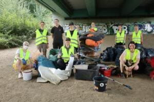 Eine Gruppe junger Erwachsener in Warnwesten posiert mit einem Hund unter einer Brücke neben großen Mengen gesammelten Mülls, darunter Styropor, Säcke und ein Einkaufswagen.