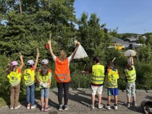 Grundschulkinder und eine Lehrerin in Warnwesten recken von hinten fotografiert Stöcke und gefüllte Beutel in die Luft.
