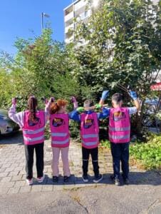 Vier Kinder in pinken World-Cleanup-Day-Westen stehen von hinten fotografiert vor Büschen und heben die Hände mit Handschuhen.