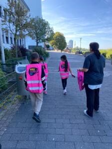 Kinder in pinken World-Cleanup-Day-Westen gehen mit einer Lehrerin eine Uferpromenade entlang, von hinten fotografiert.