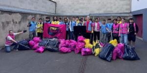Eine große Gruppe Jugendlicher in Warnwesten steht vor einer Betonwand mit pinker World-Cleanup-Day-Fahne, davor viele gefüllte Müllsäcke und ein Gitter.