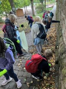 Schulkinder mit Rucksäcken, Handschuhen und Greifzangen sammeln Müll im Laub entlang einer alten Steinmauer.