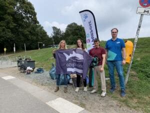 Vier Erwachsene halten eine Fahne des Vereins Ozeankind an einem Deich bei einem Radweg, daneben Beachflag, Lastenrad und gefüllte Müllsäcke.