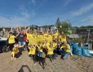 Eine große Gruppe in gelben Trash-Hero-T-Shirts jubelt mit erhobenen Armen hinter einem Banner, daneben ein Hund und viele gefüllte blaue Müllsäcke.