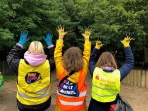 Drei Kinder in Warnwesten heben von hinten fotografiert ihre Hände in bunten World-Cleanup-Day-Handschuhen in die Luft.