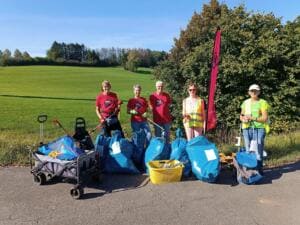Fünf Erwachsene, einige in roten World-Cleanup-Day-T-Shirts, stehen mit Greifzangen und Handwagen hinter blauen Müllsäcken am Rand einer Wiese, daneben eine pinke Beachflag.