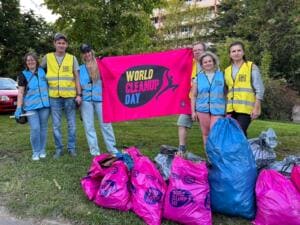 Sechs Erwachsene in blauen und gelben Warnwesten halten eine pinke World-Cleanup-Day-Fahne, davor viele gefüllte Müllsäcke auf einer Wiese.