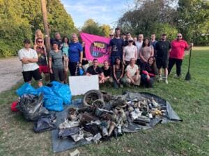 Eine große Gruppe steht hinter einer pinken World-Cleanup-Day-Fahne, davor auf einer Plane viele aus dem Wasser geborgene Fundstücke wie Reifen, Flaschen und Metallteile; eine Person hält ein Stand-up-Paddel.