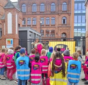 Viele Kinder in bunten World-Cleanup-Day-Westen stehen vor dem Tor einer Backstein-Schule, an dem eine gelbe World-Cleanup-Day-Fahne hängt.