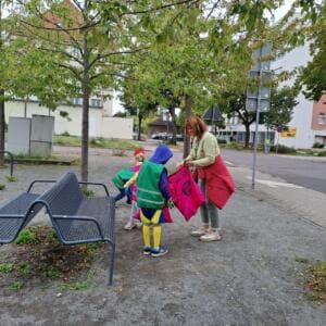Kleine Kinder in grünen Westen sammeln mit einer Erzieherin und pinken Säcken Müll an einer Bank am Gehweg.