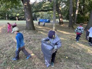 Kinder mit großen grauen Müllsäcken sammeln Müll auf einer Wiese unter alten Bäumen in einem Park.