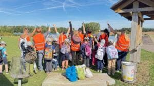 Eine Gruppe Kinder mit Rucksäcken und Erwachsene in orangefarbenen Warnwesten steht von hinten fotografiert am Seeufer und hebt die Hände.